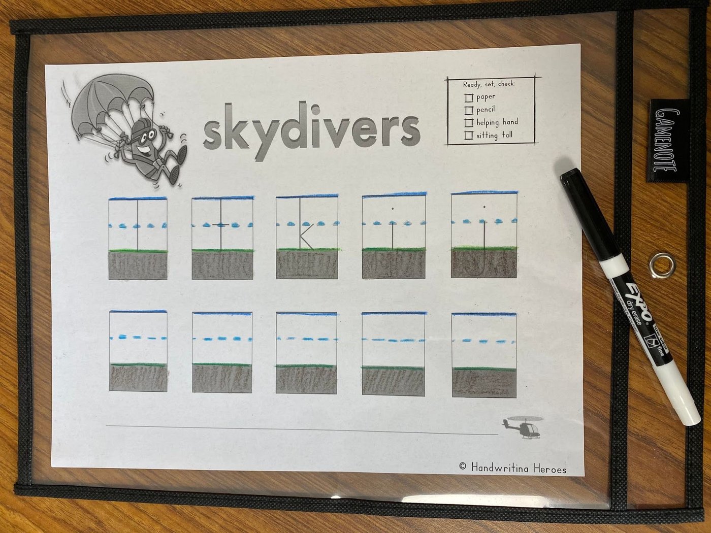 A Handwriting Heroes skydivers worksheet inside a dry erase sleeve on a classroom desk, with the sky, cloud, grass and dirt lines colored in by hand and an Expo marker beside it
