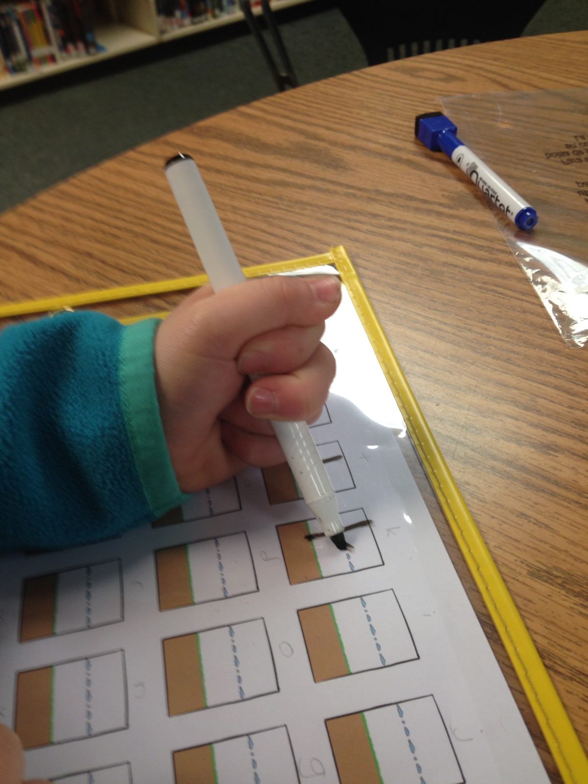A child's hand holding a dry-erase marker in a fisted thumb-wrap grasp over a letter practice sheet