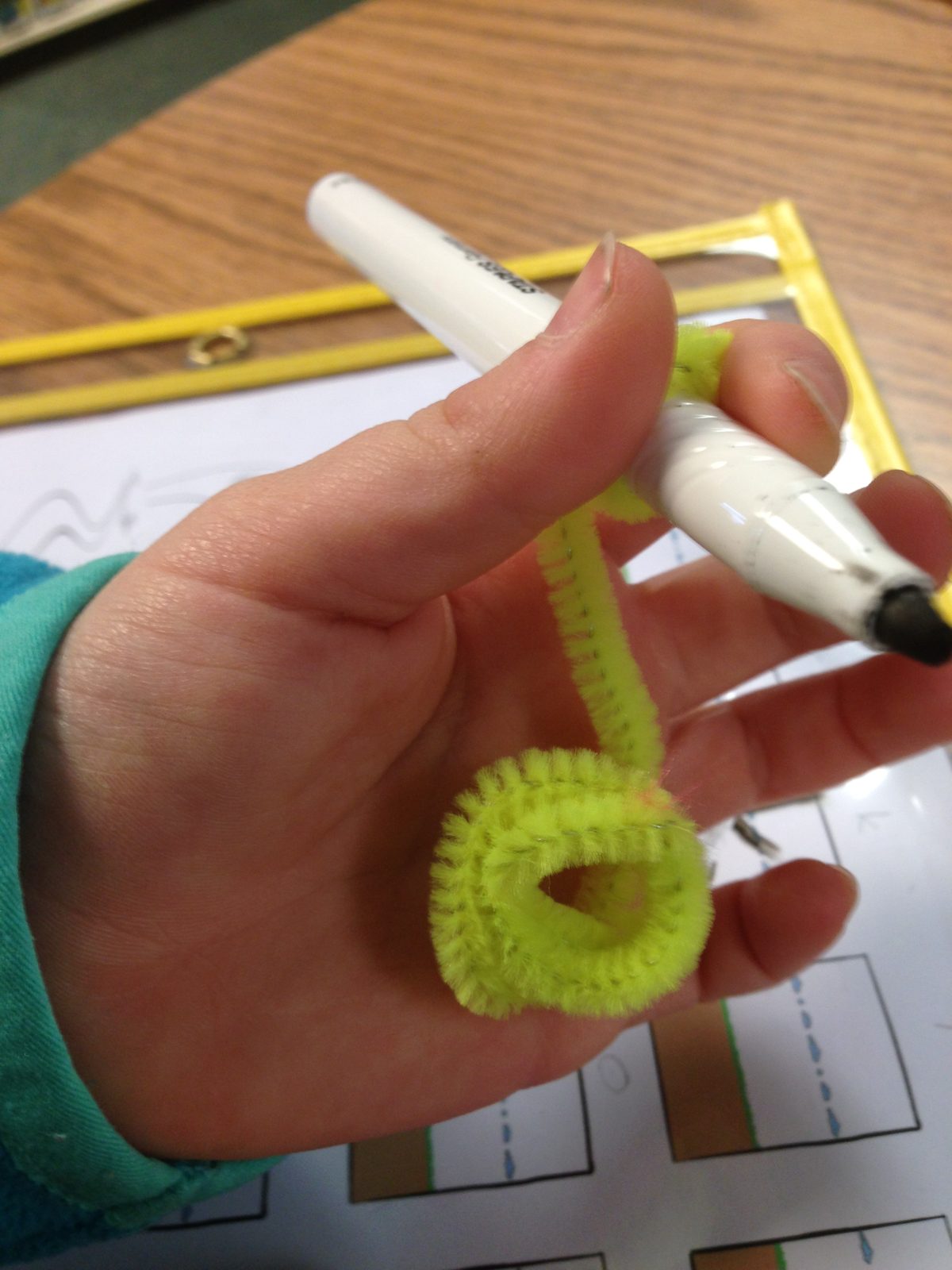A child's hand holding a coiled yellow Wikki Stix against the palm with the pinkie and ring fingers, with the other end wrapped around a marker