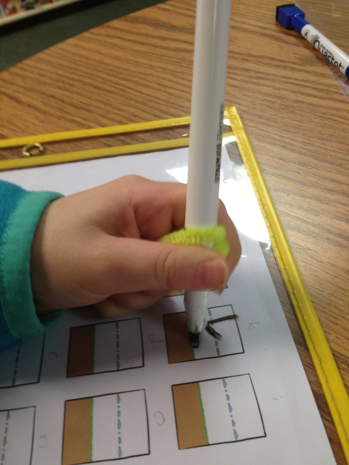 The same child writing on a letter practice sheet, now holding the marker in a tripod grasp with the yellow Wikki Stix wrap at the grip point