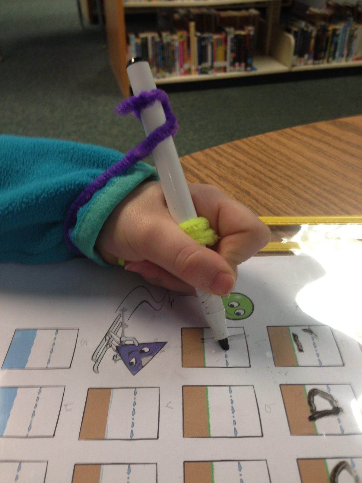 A child writing with a marker that has a purple Wikki Stix connecting its top to the wrist, guiding the marker to rest in the web space of the hand